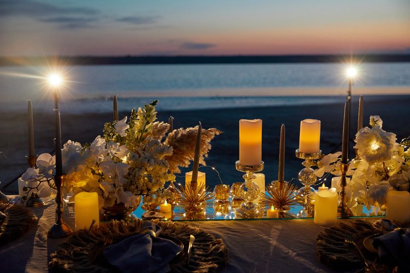 Elegant evening table setup by a serene shoreline at sunset, featuring an array of glowing candles in varying heights. Beautiful white and cream floral arrangements adorn the table, alongside decorative elements like metallic starburst accents. The centerpiece stands on a reflective surface amplifying the ambient candlelight. The background showcases a gently rippling body of water under a fading twilight sky. The cozy, intimate atmosphere is enhanced by the warm, soft lighting and the tranquil natural setting.