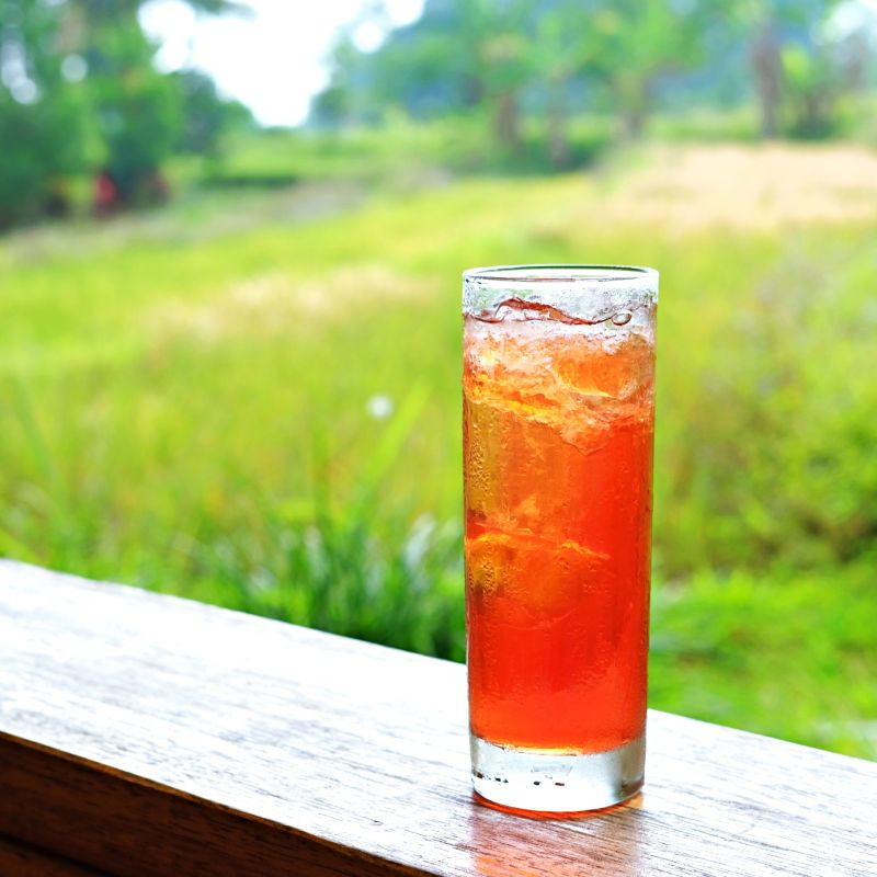 A refreshing glass of iced red tea sits on a wooden ledge overlooking a lush green field. The bright,vibrant color of the tea contrasts with the serene natural backdrop, perfect for a relaxing outdoor