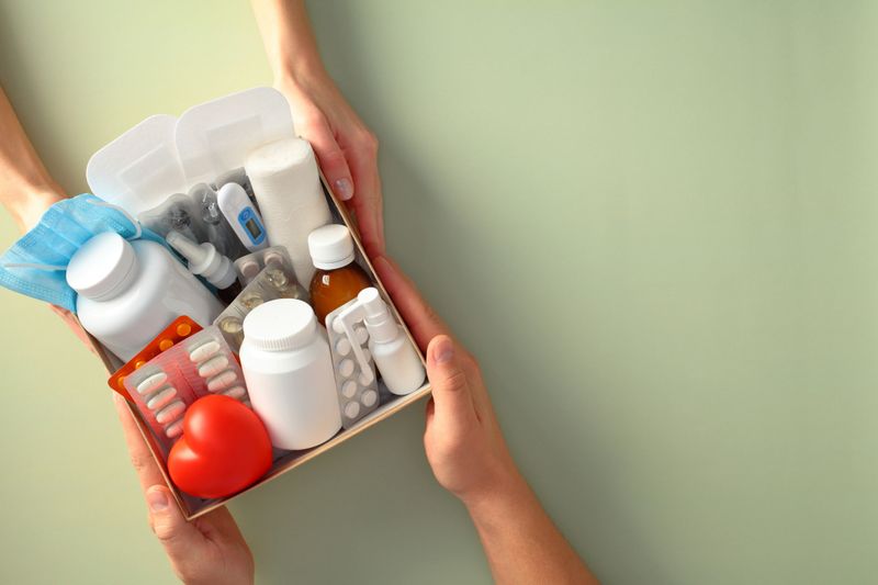 Hands holding a first aid kit containing various medical supplies, including pills, a thermometer, bandages, and a face mask, against a green background