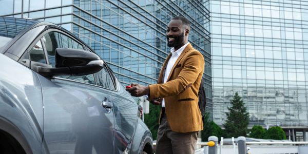 A man in a brown jacket smiling while unlocking his car outside a glass building.