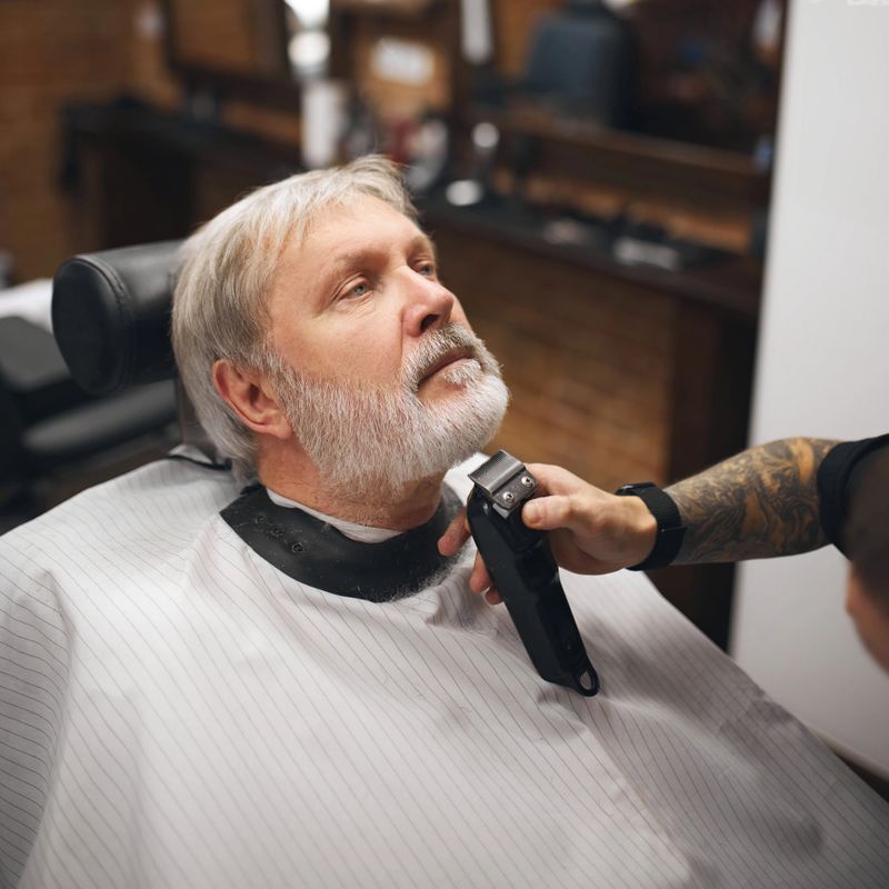 Barber carefully using clipper to finish detailing client beard, focusing the contours of chin. Elderly man visiting barber shop Concept of male beauty, barbershop, hairstyle, salon, hairdresser