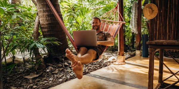 Man relaxing in a hammock using a laptop surrounded by greenery.