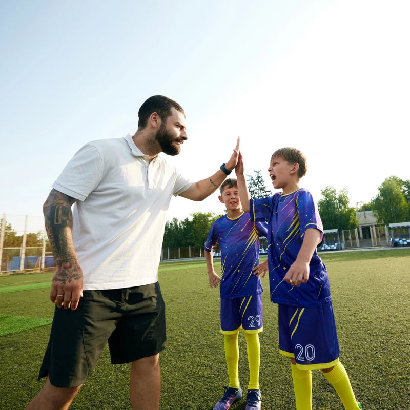 Bearded man, soccer coach giving high-five to kids in purple uniform, supporting children in the outdoor game session. Concept of sport, childhood, education, achievement, active lifestyle