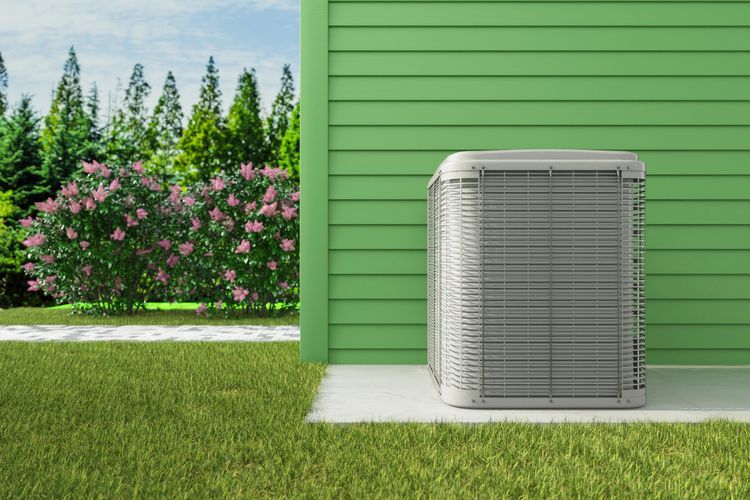 Outdoor air conditioning unit next to a green house with flowers and trees in the background.