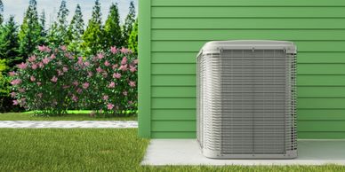 Outdoor air conditioning unit next to a green house with flowers and trees in the background.