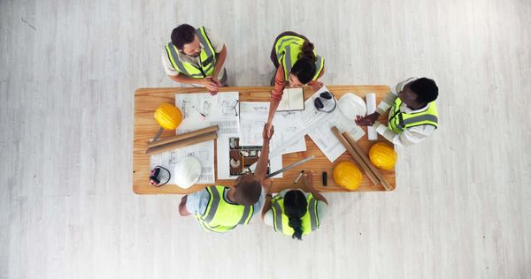 Engineers in safety vests shaking hands over architectural plans on a wooden table.