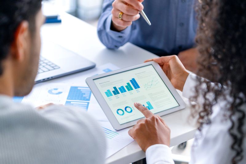 Close up of three people looking at financial data with graphs and charts.  All their hands can be seen and one person is pointing with a pen. There is paperwork on the desk showing more finance information