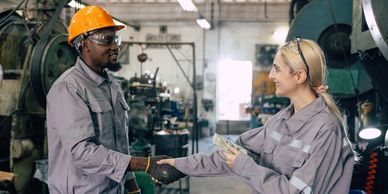 Two factory workers shaking hands, one holding money, in an industrial setting.