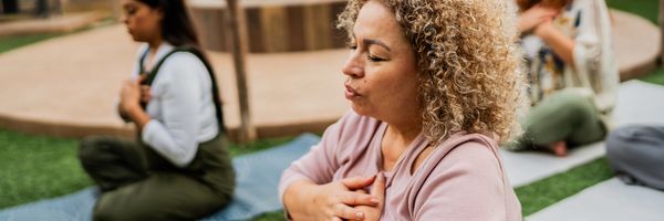 Women meditating outdoors with hands on chest near a fire pit.