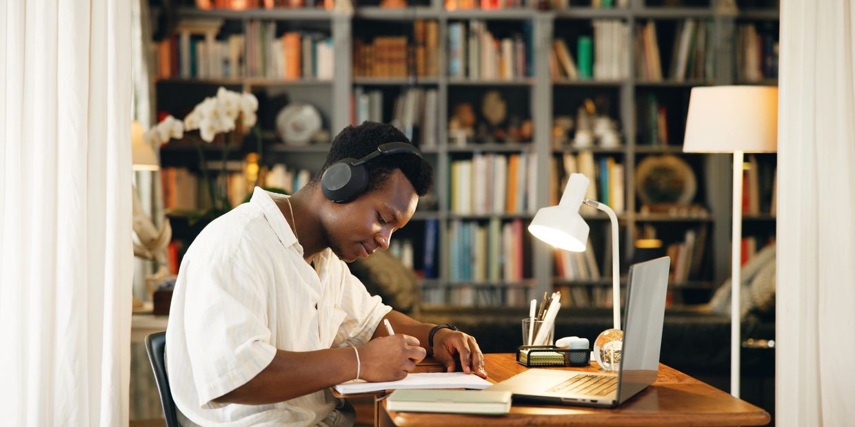 A man wearing headphones writes in a notebook at a desk with a laptop in a cozy library setting.