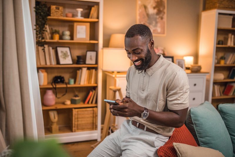 A smiling black man is sitting on the back of a sofa and using a mobile phone to send text messages