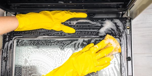 Hands in yellow gloves cleaning oven door with sponge.