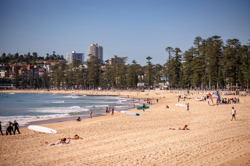 Warm sunlight bathes Manly Beach as locals and visitors revel in a perfect day by the ocean. Surfers catch waves while sunbathers lounge on the soft sand, creating a lively atmosphere.