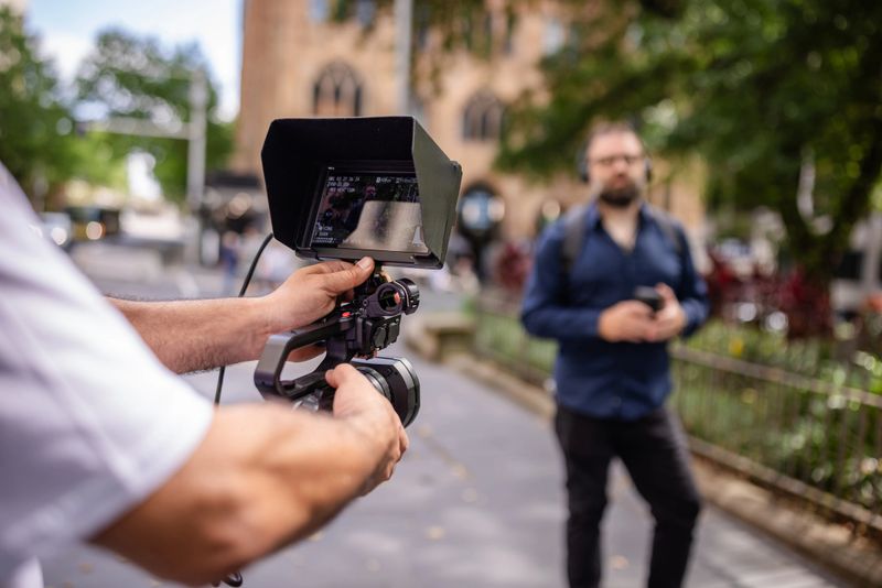 A videographer focuses on a man standing in a busy area of Sydney, Australia. The scene showcases urban life with pedestrians and lush greenery in the background.