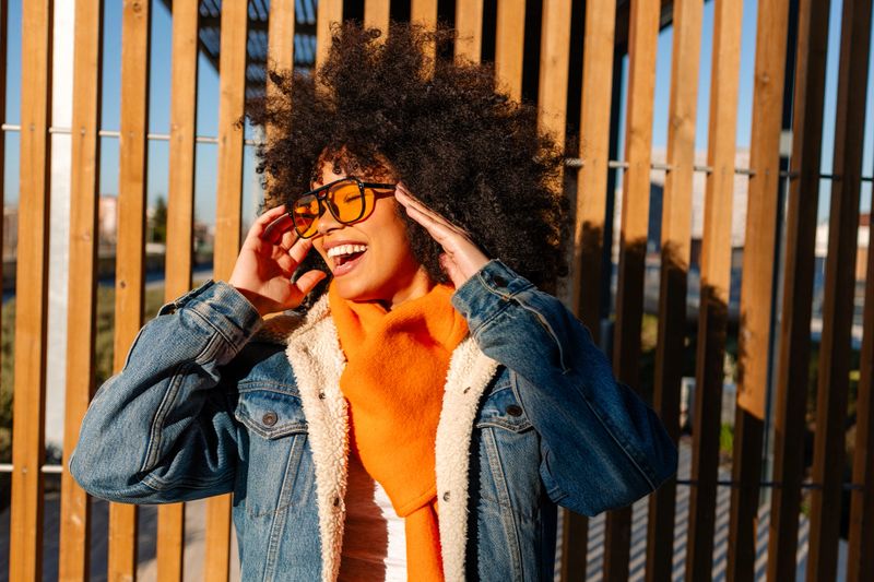 Stylish young woman with afro hair, wearing orange scarf and denim jacket, smiling and adjusting sunglasses against wooden background