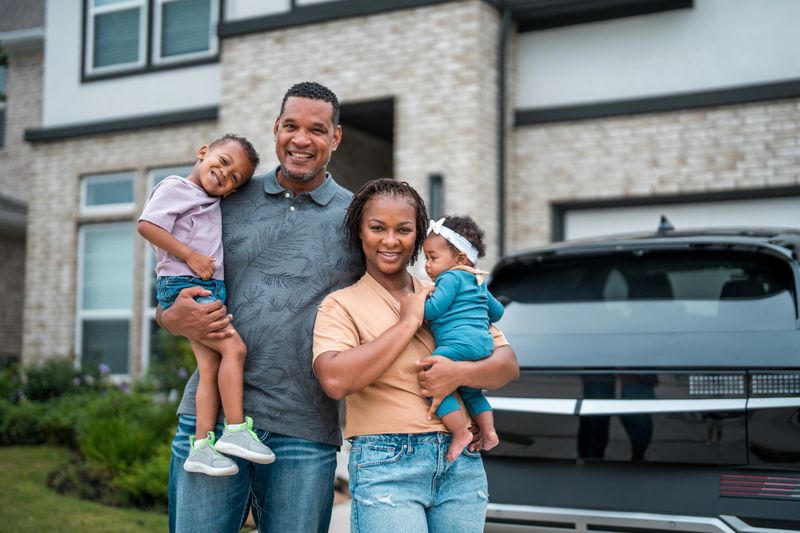 A cheerful diverse family stands outside their solar-powered home, with a man and two children smiling near an electric vehicle, signifying sustainable lifestyle choices.