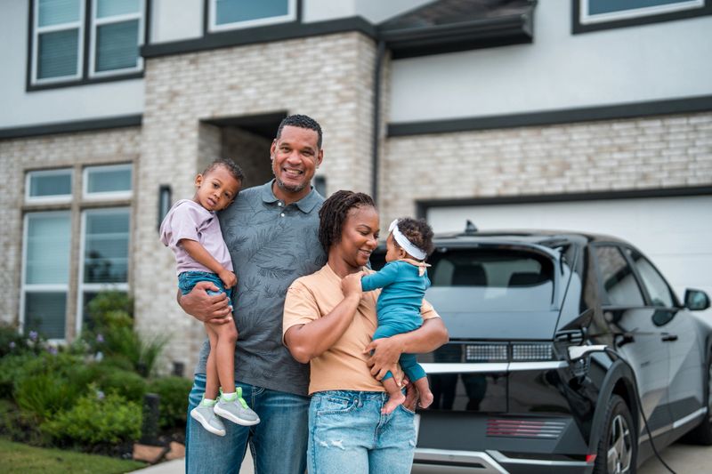 A smiling diverse family enjoying time together outside their solar-powered home, with an electric vehicle and grocery bags nearby, symbolizing sustainable lifestyle choices.