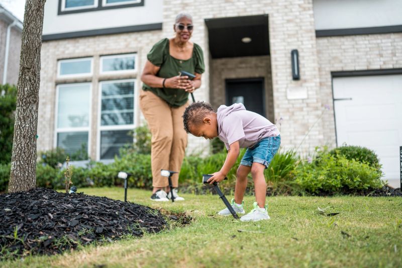 A young mixed-race child and his smiling grandmother install solar lights on a sunny day, exemplifying sustainable living while enhancing their home's exterior.