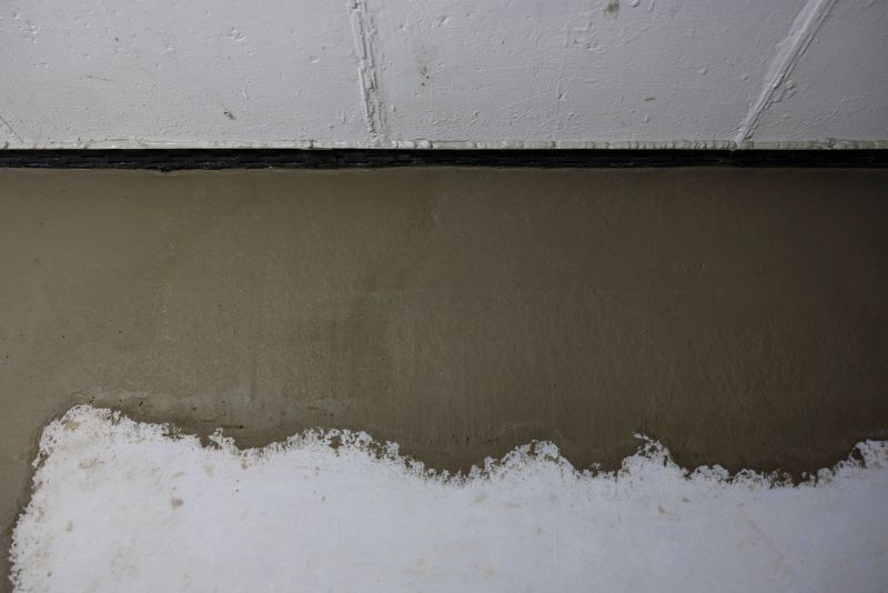 Fresh concrete-based protective coating covers upper portion of basement wall creating distinct contrast against untreated white surface below. Specialized waterproof barrier application demonstrates professional foundation treatment process for moisture protection