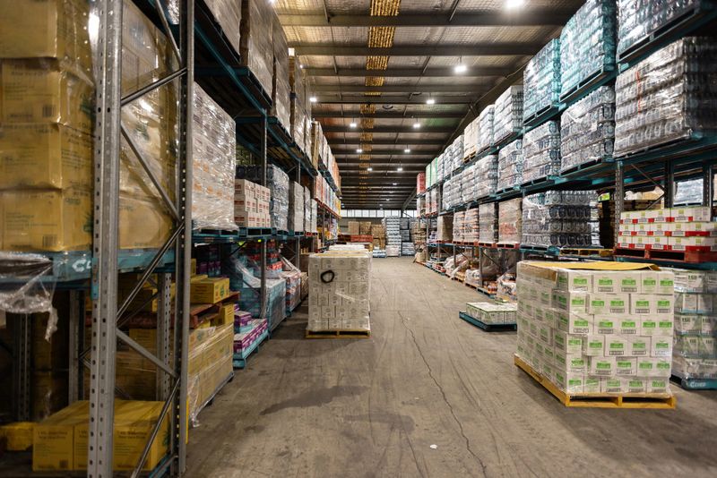 View of a spacious warehouse interior featuring neatly stacked goods on industrial shelves with organized pallets on the floor under bright artificial lighting, showcasing efficient storage organization and distribution logistics.