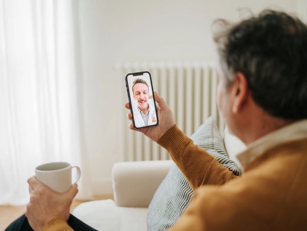 Older man video calling a doctor from home, holding a cup.