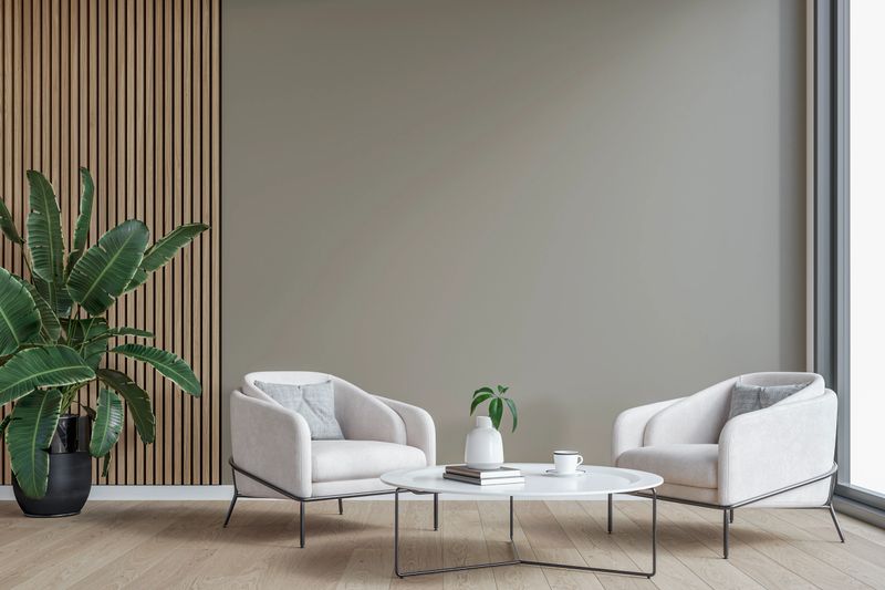 This image showcases a modern, minimalist seating area with two elegant white armchairs positioned around a sleek, low-profile white coffee table in front of a light gray blank plaster wall and copy space. A wooden slat accent wall on one side adds warmth and texture,  complemented by a large green plant in a black pot. Natural light from the floor-to-ceiling window enhances the soft, neutral tones, creating a bright and inviting atmosphere. 3D rendered image.