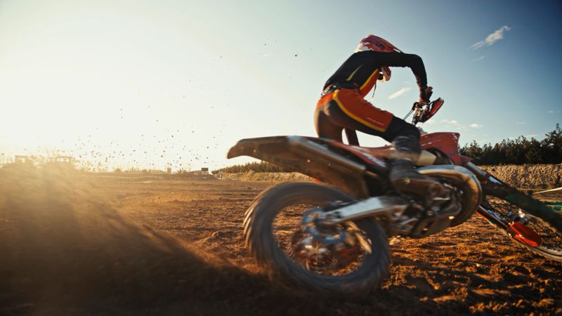 A Caucasian female in racing gear speeds across a dirt motocross track at sunset, showcasing energy and skill. The movement of the bike and flying dirt adds dynamic excitement.
