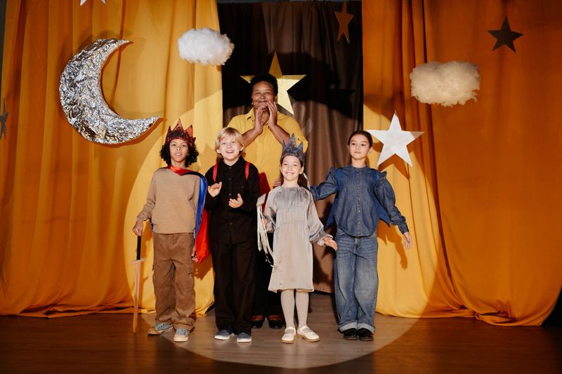 Children dressed in costumes standing on stage during school play with orange curtain background depicting night sky elements including moon and stars