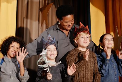 Children wearing star crowns and a woman smiling behind them on stage.