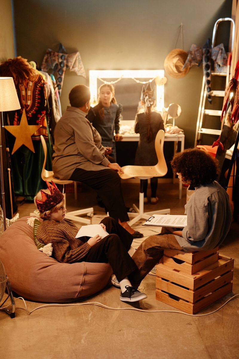 Group of children sitting and standing in dressing room preparing for play, with wigs, costumes, and script on hand, engaging in theater activities