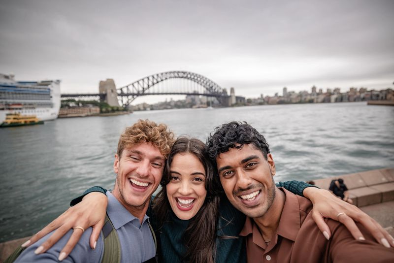 A group of young adults stands by the water at Sydney Harbour, smiling widely as they embrace and take a cheerful selfie. The iconic bridge and city skyline create a lively backdrop.