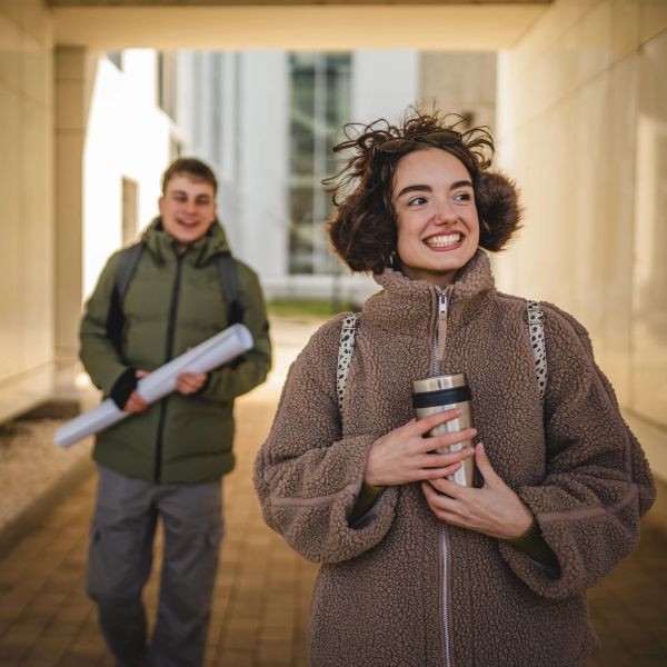 Happy woman with earmuffs holding a travel mug, man in background with rolled papers.