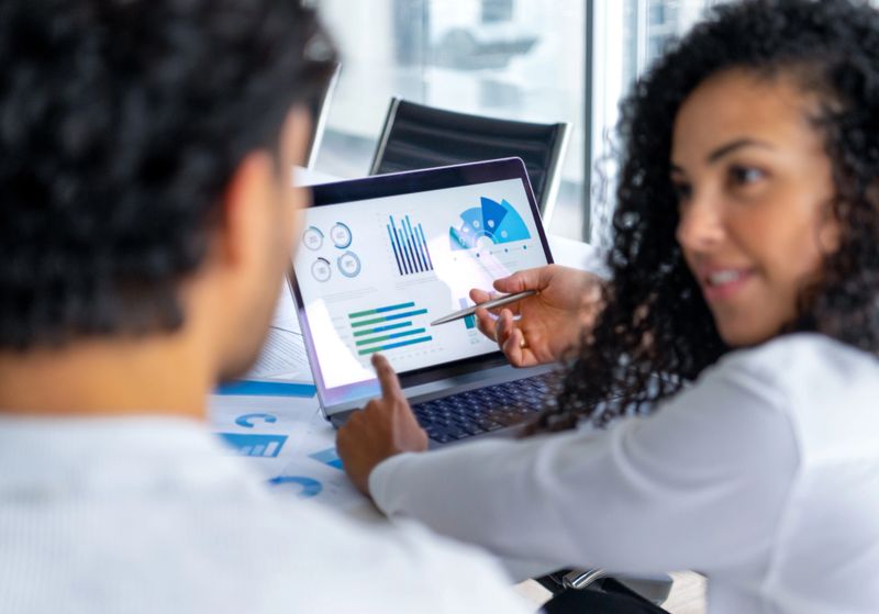 A couple looking at and discussing  financial data with graphs and charts.  A business man and woman can be seen and one person is pointing at a laptop computer. There is paperwork on the desk showing more finance information