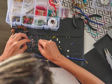 Person crafting a beaded necklace at a workspace with tools and beads.