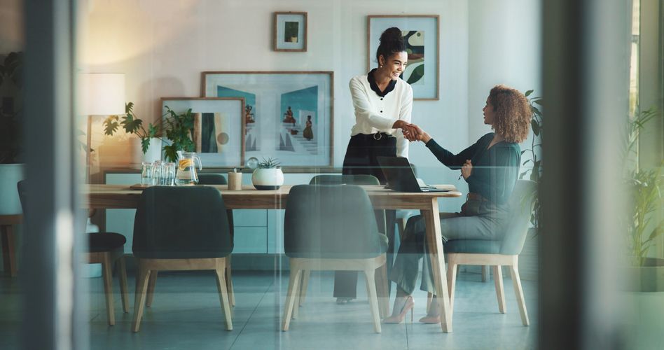 Two women shaking hands in a modern office meeting room.