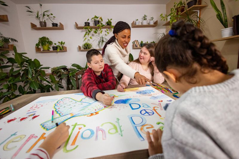 A multicultural classroom gathers around a giant paper, enthusiastically drawing and painting a dove for International Day of Peace, guided by their caring teacher.