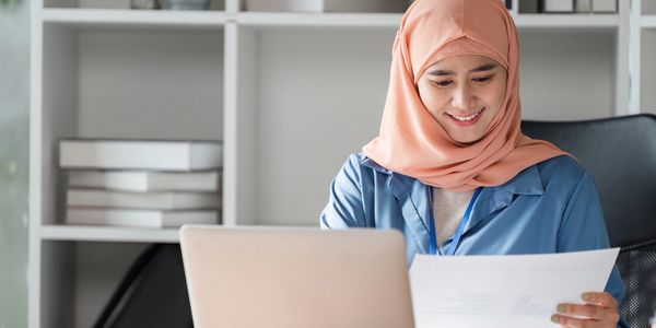 A woman in a peach hijab smiles while reviewing documents at her desk.
