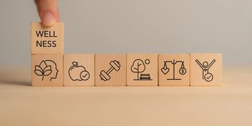 Wooden blocks with wellness symbols and a finger placing the word 'WELLNESS'.