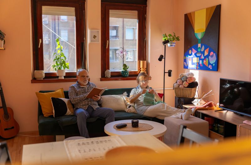 A serene living room scene featuring an elderly couple relaxing, with one engaged in knitting and the other reading a book, surrounded by cozy decor and bright natural light.