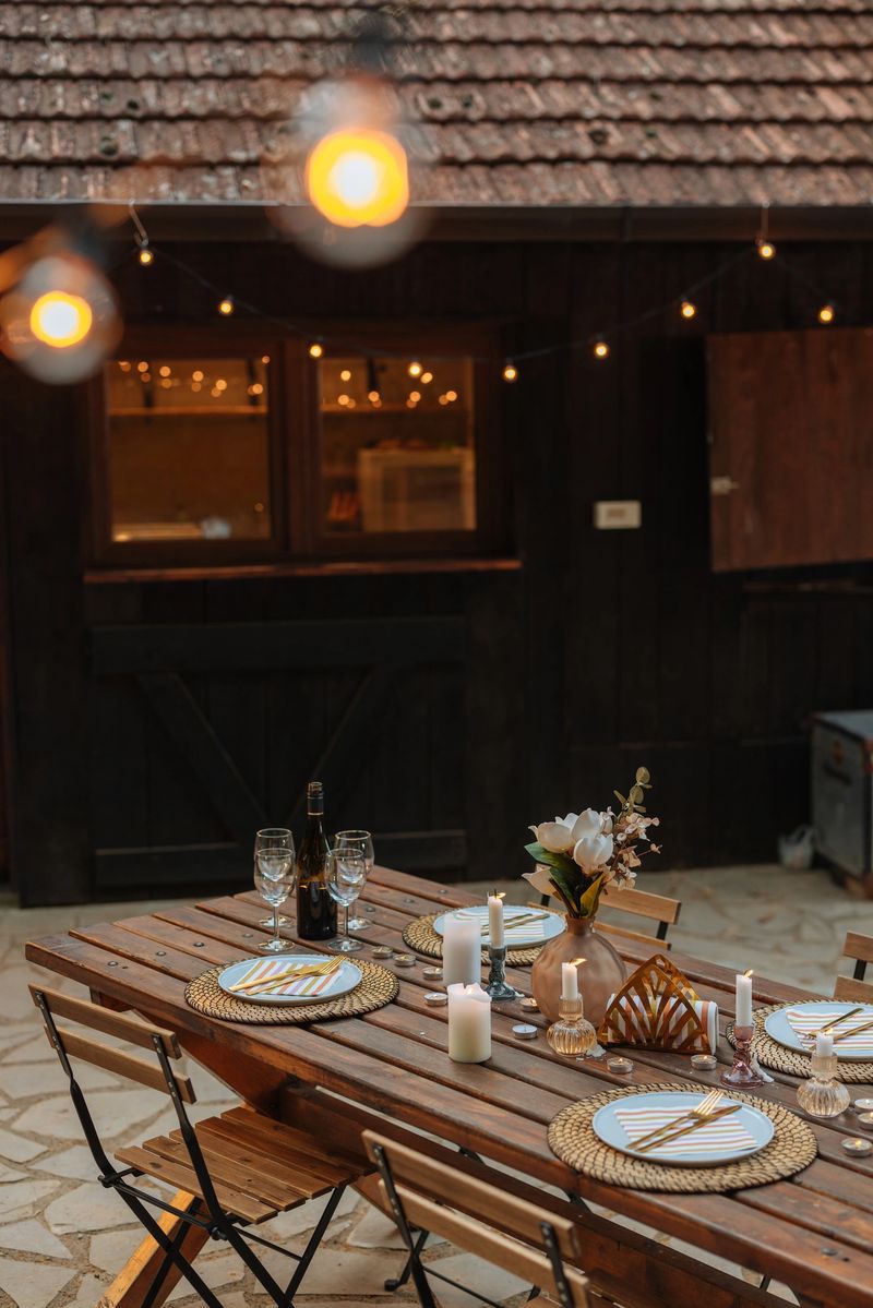 Charming outdoor dining area with a wooden table adorned with plates, glasses, and candles, enhanced by warm string lights