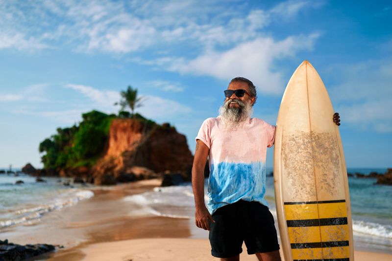 Elderly man with surfboard on sunny beach. Active senior enjoying ocean view, healthy retirement lifestyle adventure. Surfing as leisure in golden years. Celebrating vitality new experiences.
