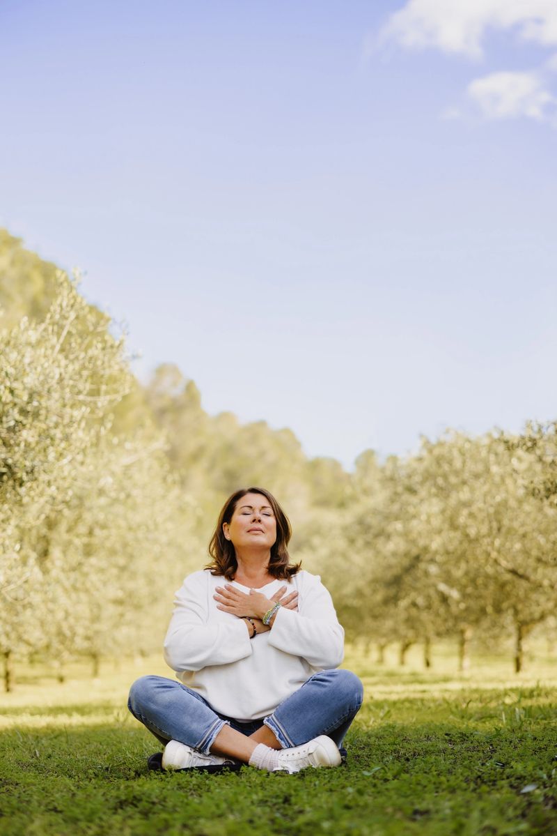 Mature woman in her menopausal years sitting in a cross-legged position surrounded by delicate young olive trees with her hands placed over her heart in a mindfulness pose. She embraces the stillness of the moment, radiating tranquility and self-awareness. Color editing. Part of a series.