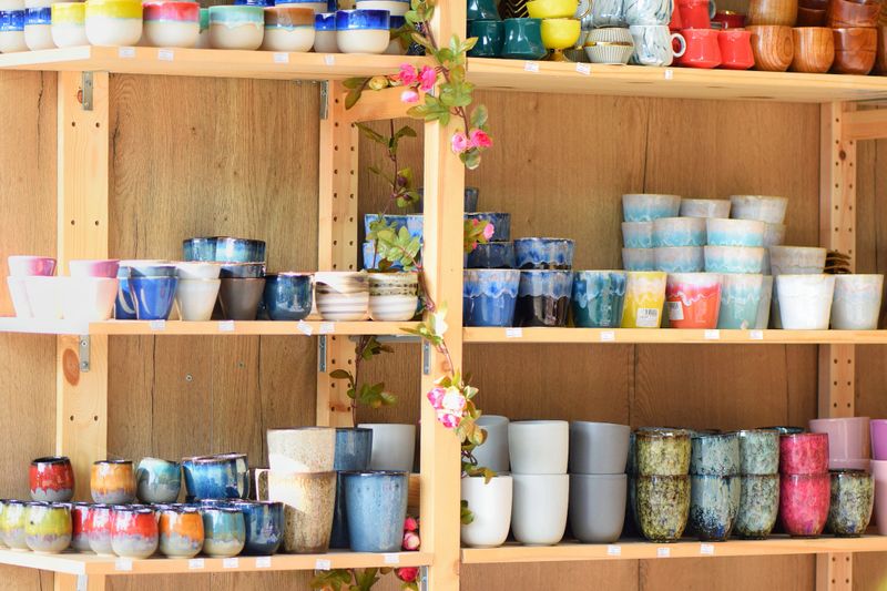 Various handmade ceramic cups and small pots arranged on wooden shelves, surrounded by decorative artificial flowers. A vibrant mix of colors, textures, and glaze patterns, ideal for showcasing artisan crafts or home decor.
