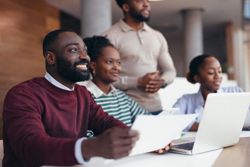 Group of young African businesspeople participating in a meeting within a modern office, smiling and engaging in conversation while looking towards each other and their laptops