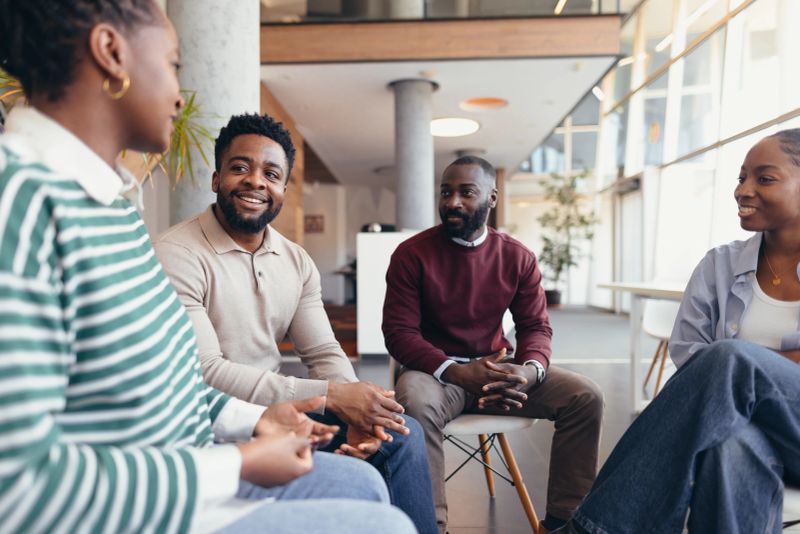 Four young professionals are sitting in a circle in a modern office, having a discussion and sharing ideas, fostering teamwork and collaboration