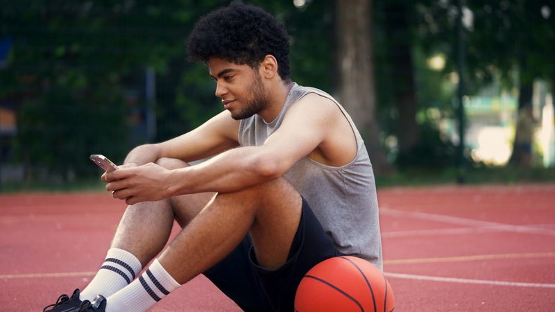 Young man sits on the basketball court, scrolling on his smartphone and using a sports betting app