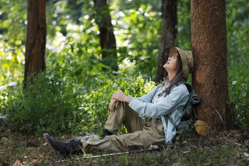 A female hiker sits under a tree in a lush forest, resting and enjoying the peaceful surroundings. Wearing a hat and outdoor gear, she looks up at the trees, reflecting on nature's beauty and tranquility