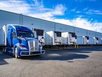 Blue semi truck parked in front of a warehouse with several docked trailers.