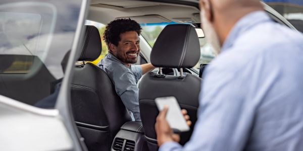 Man smiling while sitting in driver's seat of car talking to another man.