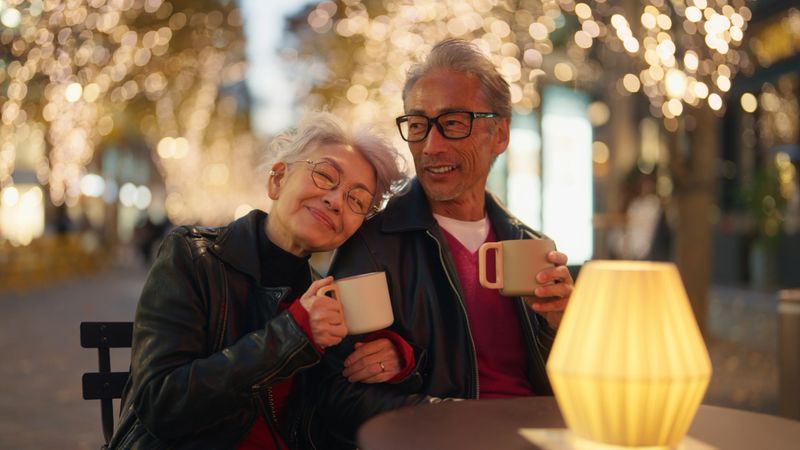 A senior couple sits closely together at a cozy outdoor café. The woman leans her head lovingly on her partner's shoulder while holding a warm mug. Surrounded by twinkling festive lights, they share a serene moment of connection and relaxation, enjoying the enchanting evening ambiance.